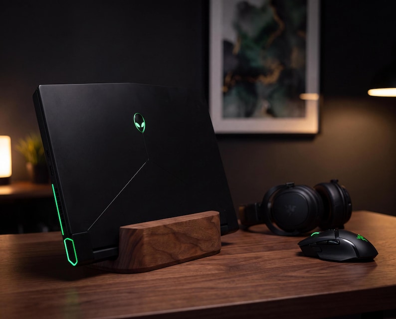 a sleek black laptop with a green alien logo on the back, placed on a wooden desk. The laptop is accompanied by a mouse and headphones, suggesting a workspace setup for gaming or multimedia purposes.