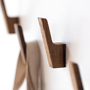 May include: A series of wooden wall hooks with a minimalist design. The hooks are a warm brown color and are mounted on a white wall. The hooks have a simple, angular shape, perfect for hanging coats, bags, or other items.