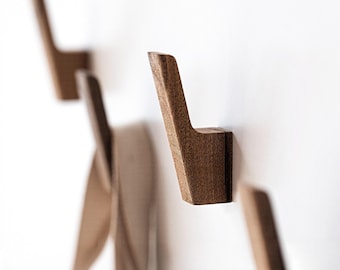 May include: A series of wooden wall hooks with a minimalist design. The hooks are a warm brown color and are mounted on a white wall. The hooks have a simple, angular shape, perfect for hanging coats, bags, or other items.