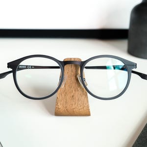 A pair of black-framed glasses is placed on a wooden stand on a white surface, with a black vase and a black object in the background.