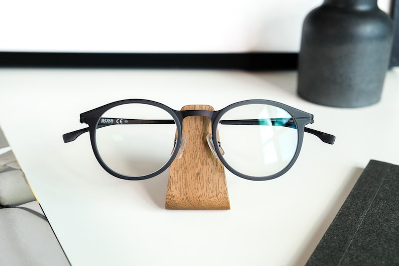 A pair of black-framed glasses is placed on a wooden stand on a white surface, with a black vase and a black object in the background.