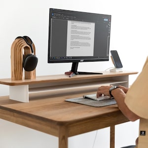 May include: A wooden desk with a monitor, keyboard, and a wooden headphone stand. The desk has a light wood finish and a white support structure. A pair of black headphones hangs on the stand. A smartphone sits on a wireless charger.