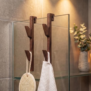 a bathroom setting with a glass shelf displaying two wooden towel hooks, a white towel, and a glass vase with greenery.