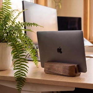 a wooden stand holding a laptop, with a potted plant and a computer monitor in the background.