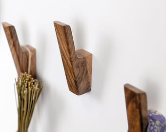 May include: Three wooden wall hooks with a natural wood grain finish are mounted on a white wall. The hooks have a modern, angular design, and one hook holds a bundle of dried plants. The hooks are designed for hanging items.