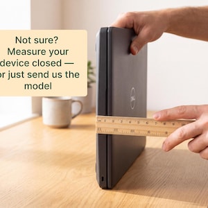 A person's hands are holding a ruler and measuring the size of a closed laptop computer on a wooden desk.