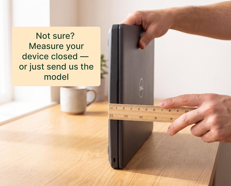 A person's hands are holding a ruler and measuring the size of a closed laptop computer on a wooden desk.