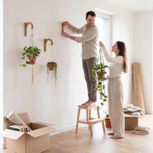 A man and a woman are hanging plants from wooden hooks on a wall in a room with a cardboard box and some books.