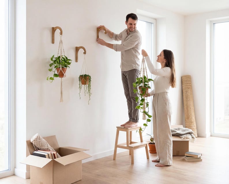 A man and a woman are hanging plants from wooden hooks on a wall in a room with a cardboard box and some books.
