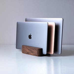Puede incluir: Un soporte vertical para portátil de madera marrón oscuro que sostiene tres portátiles en fila. El portátil delantero es plateado con un logotipo de Apple negro, el del medio es oro rosa y el trasero es gris espacial. El soporte tiene forma redondeada.