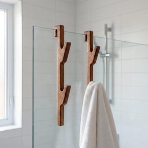 Könnte beinhalten: Zwei hölzerne Duschhaken mit einem Astdesign. Ein Haken hält ein weißes Handtuch. Die Haken sind aus dunkelbraunem Holz gefertigt und hängen an einer Glas-Duschabtür. Der Hintergrund ist eine weiße geflieste Wand.