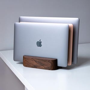 Pode incluir: Um suporte de madeira para laptop com três laptops prateados e ouro rosa. O suporte é feito de madeira escura e tem um design curvo. O logotipo da Apple é visível na frente do laptop.