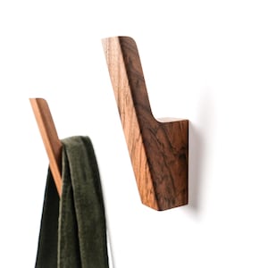 Puede incluir: Tres ganchos de madera montados en la pared con un acabado de veta de madera marrón oscuro. Cada gancho presenta un diseño en ángulo que apunta hacia arriba. Un gancho sostiene una prenda de tela verde oscuro, mientras los otros están vacíos.