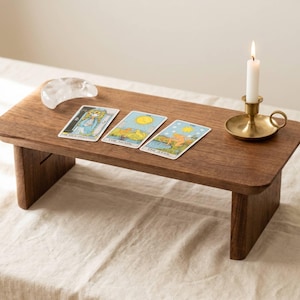 Peut inclure: Petite table rectangulaire en bois aux coins arrondis. Trois cartes de tarot, un cristal clair et une bougie allumée dans un bougeoir en laiton sont disposés sur la table. Les cartes sont intitulées "The High Priestess", "The Moon" et "The Star".