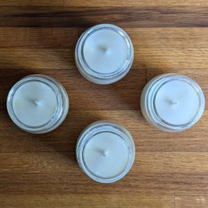 Peut inclure: Quatre bougies blanches dans des pots en verre avec une seule m&egrave;che dans chacun. Les bougies sont sur une surface en bois.