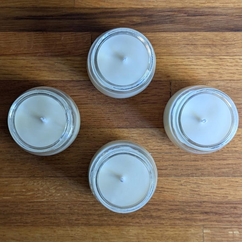 Peut inclure: Quatre bougies blanches dans des pots en verre avec une seule m&egrave;che dans chacun. Les bougies sont sur une surface en bois.