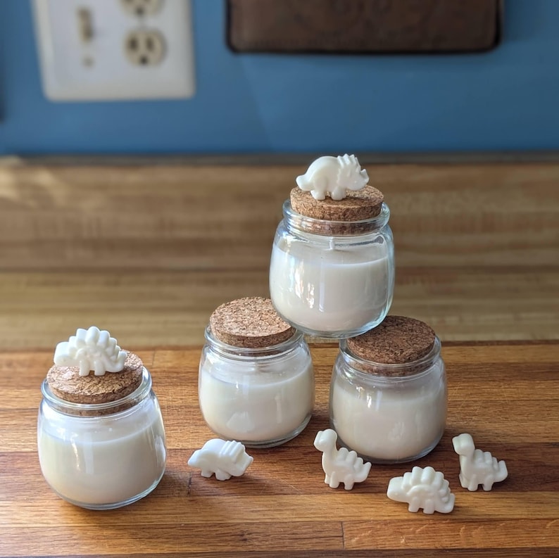 Peut inclure: Quatre petits pots en verre avec des couvercles en li&egrave;ge et des bougies blanches. Chaque pot est surmont&eacute; d'une petite figurine de dinosaure blanche. Les pots sont dispos&eacute;s en forme de pyramide sur une surface en bois.