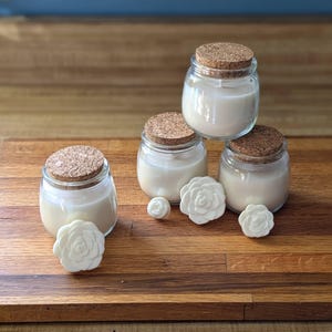 Peut inclure: Quatre pots en verre avec des couvercles en li&egrave;ge remplis de bougies en cire blanche. Les bougies sont dispos&eacute;es en forme de pyramide sur une surface en bois. Il y a quatre d&eacute;corations de roses en c&eacute;ramique blanche sur la surface.