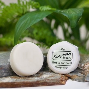 May include: Two round, white shampoo bars are displayed on grey and brown stones, with green foliage behind them. One bar is labelled with "Koopman Soap Co" and "Lime & Patchouli Shampoo Bar". The label also states the bar weighs 50g.