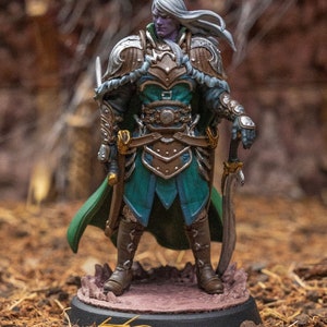 May include: A painted miniature figurine of a warrior with long white hair, wearing a green and gold armor with a sword and dagger. The figurine is standing on a round base.