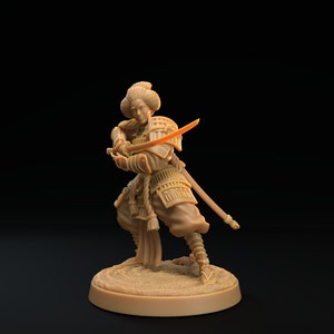 May include: A brown painted miniature figurine of a samurai warrior holding a sword. The figurine is standing on a round base.