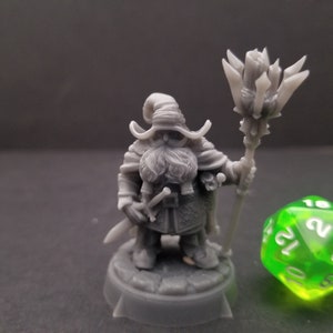 May include: A grey 3D printed miniature of a dwarf wizard holding a staff with a spiky top. The dwarf is wearing a hat and a long beard. The miniature is standing on a round base.
