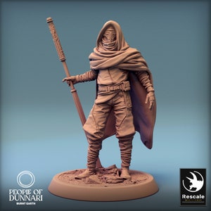 May include: A painted miniature figurine of a hooded figure holding a staff. The figure is wearing a long robe and has a mask over their face. The miniature is painted in shades of brown and beige.