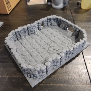 May include: A grey, stone-textured, rectangular gaming tile with a raised border. The tile has a grid pattern and is designed to represent a dungeon or cave setting.
