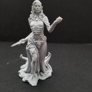 Peut inclure: Une miniature en plastique gris imprimée en 3D d'une figure féminine aux cheveux longs, portant une robe fluide et tenant un petit objet dans sa main droite. Elle se tient sur une base avec un motif tourbillonnant.
