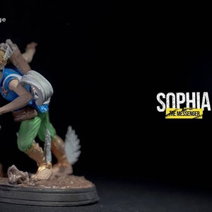 Puede incluir: Una figura miniatura pintada de un personaje femenino con cabello rubio, vistiendo un atuendo azul y verde, sosteniendo un bastón. La figura está de pie sobre una base negra con una superficie texturizada marrón. El texto "Sophia The Messenger" es visible en el fondo.