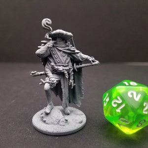 Peut inclure: Une miniature en plastique gris imprimée en 3D représentant un personnage fantastique tenant un bâton et portant une cape et un chapeau. La miniature est posée sur une base ronde. Un dé à 20 faces vert se trouve en arrière-plan.