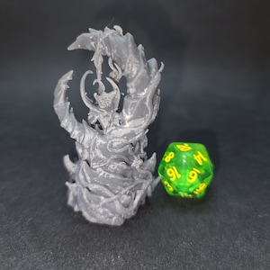 May include: A grey 3D printed miniature of a creature with a large, twisted body and a smaller figure on top. The creature is standing on a black surface next to a green 20-sided die with yellow numbers.