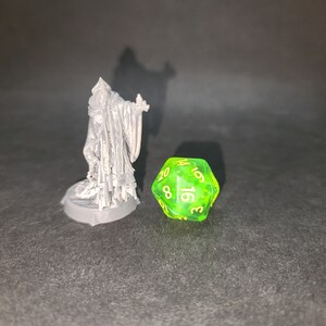 May include: A green 20-sided die with yellow numbers, next to a grey 3D printed miniature of a figure wearing a robe.
