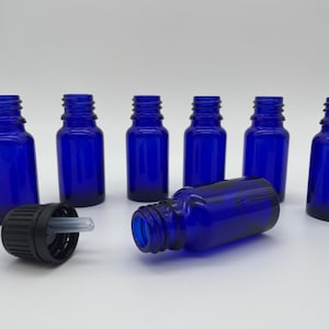 May include: Cobalt blue glass bottles with black dropper tops. The bottles are cylindrical with a rounded base and a threaded neck. One bottle lies on its side, while the others stand upright. The dropper tops are black with a clear glass dropper.