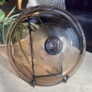 May include: A clear, brown-tinted glass lid with a central knob and a radial design. The lid is displayed on a black metal stand. The glass has a slightly reflective surface, and the overall aesthetic is vintage.
