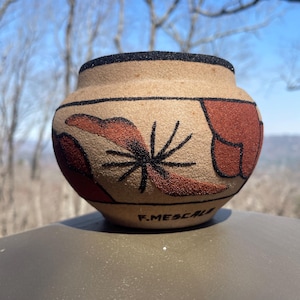 May include: A handcrafted, beige pottery vessel with a rounded shape. The pot features a black rim and is adorned with reddish-brown and black abstract designs. The artist's signature, "F. MESCALE", is visible on the base.