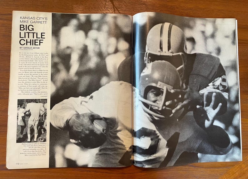 May include: A black and white magazine spread featuring the headline Kansas City's Mike Garrett Big Little Chief by Gerald Astor. The page shows a photograph of two football players in helmets and jerseys engaged in a play on the field.