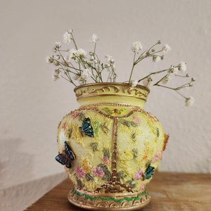 May include: A yellow ceramic vase with gold trim and floral designs. The vase is decorated with butterflies and has a small base. It is filled with white baby's breath flowers.
