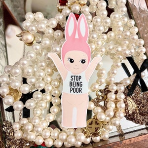 May include: A pink bunny character wearing a white tank top with the text "STOP BEING POOR" printed on it. The character is surrounded by a pearl necklace.