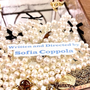 May include: A white sticker with blue text that reads "Written and Directed by Sofia Coppola" on top of a pile of white pearl necklaces and gold jewelry.
