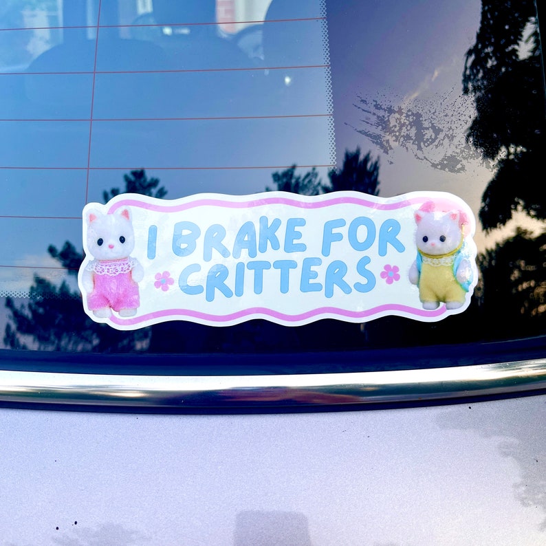 Op de afbeelding: Een roze en blauwe auto sticker met twee witte cartoon kittens. De sticker zegt "I Brake For Critters*"