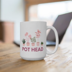 May include: White ceramic mug with a pink and green illustration of potted plants and the text "POT HEAD".