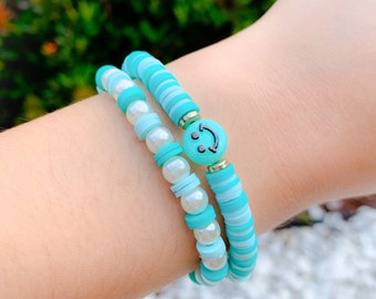 May include: Two bracelets stacked on a wrist. One bracelet features light blue and white disc beads, with a smiley face charm. The other bracelet has white pearl beads and light blue disc beads. The bracelets are a mix of light blue, white, and gold.