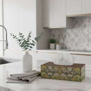 Può includere: Un portatovaglioli rettangolare con parte superiore e lati marroni, decorato con un motivo floreale nei toni del verde, giallo e crema. Una pila di tovaglioli grigi piegati e un vaso bianco con rami di eucalipto sono sul piano di lavoro.