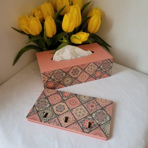 Op de afbeelding: Een roze tissueboxhoes met een kleurrijk geometrisch patroon. De hoes is gemaakt van hout en heeft een gleuf voor tissues. De doos staat op een wit oppervlak.