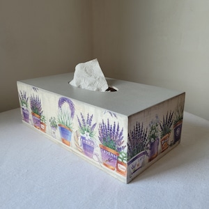 Può includere: Un portatovaglioli rettangolare con finitura verniciata grigio chiaro. La scatola è decorata con un motivo ripetuto di piante di lavanda in vasi viola e blu. Un fazzoletto bianco sporge dall'apertura ovale sulla parte superiore.