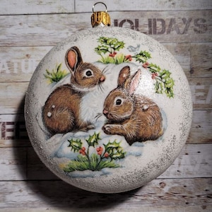 Op de afbeelding: Een ronde, witte kerstbal met een winters tafereel met twee bruine konijnen. De bal heeft een glinsterende textuur en is versierd met hulst en bessen. Bovenop zit een goudkleurige metalen lus om op te hangen.