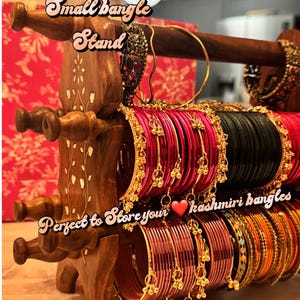 May include: A wooden bangle stand displaying various colourful bangles. The stand is brown with decorative carvings and holds rows of red, gold, and black bangles. Text on the stand reads "Pretty Shop Small bangle Stand".