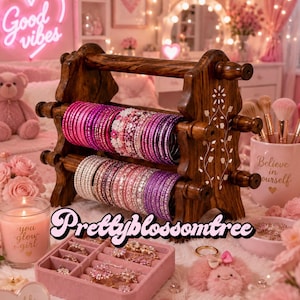 May include: A dark brown wooden jewelry stand with two horizontal bars holding rows of pink, purple, and white bangles. The stand features white floral inlay patterns on the sides. A pink velvet jewelry tray with compartments sits in front.