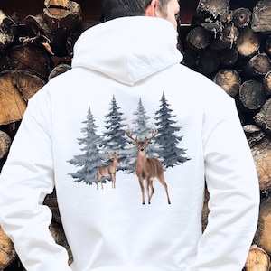 May include: A white hooded sweatshirt featuring a graphic print on the back. The design displays a large buck with antlers and a smaller deer standing in front of a cluster of dark grey evergreen pine trees against a plain white background.